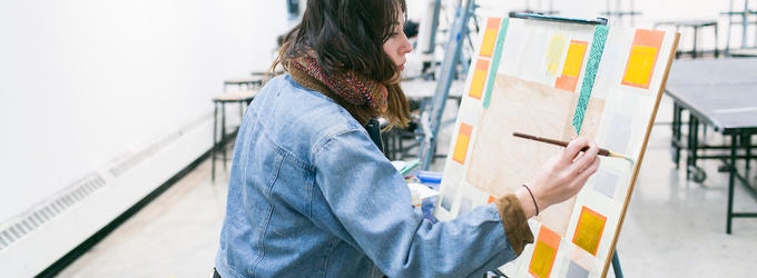 Student in a denim jacket painting at an easel